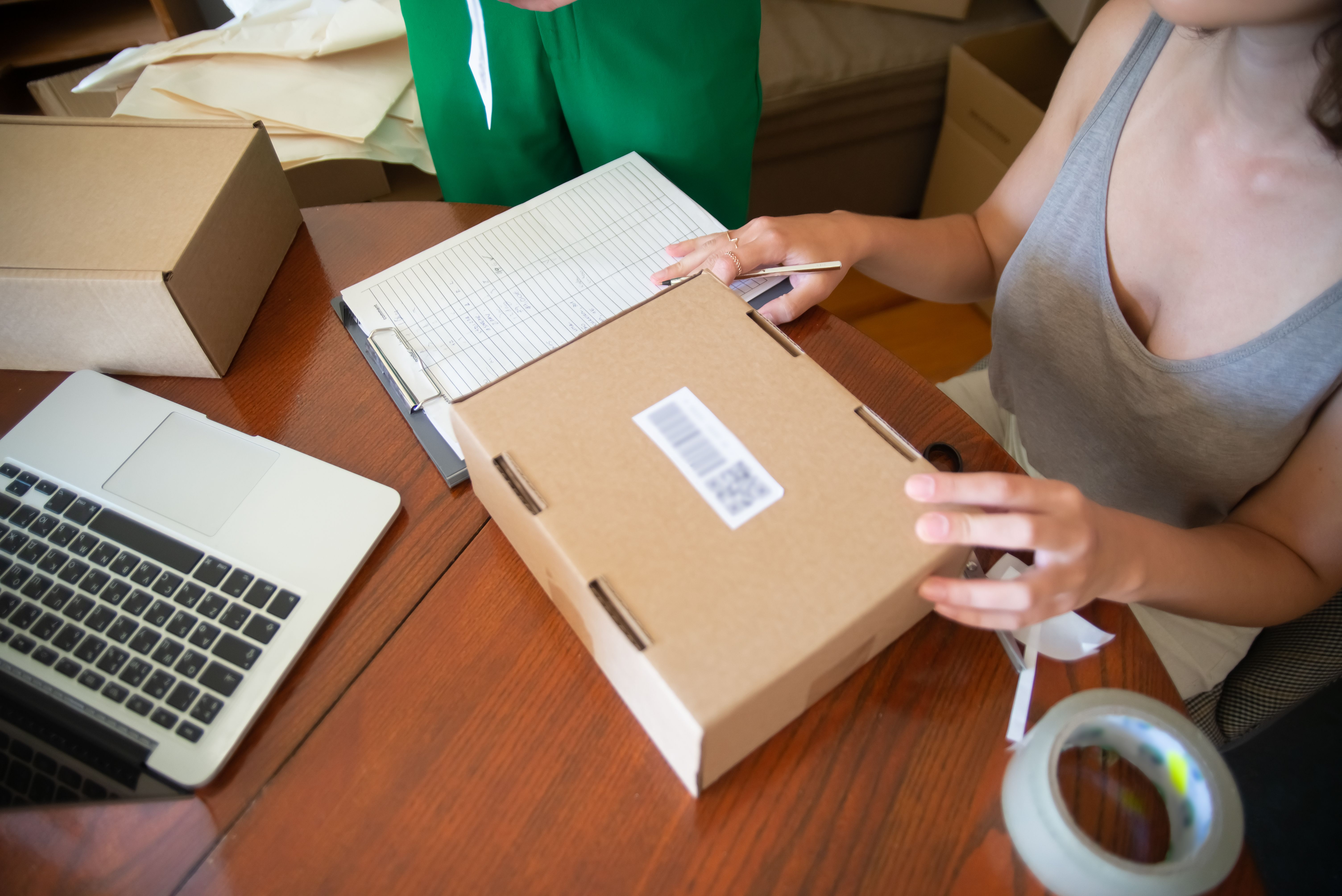 Person packing a cardboard parcel beside an order log and laptop.