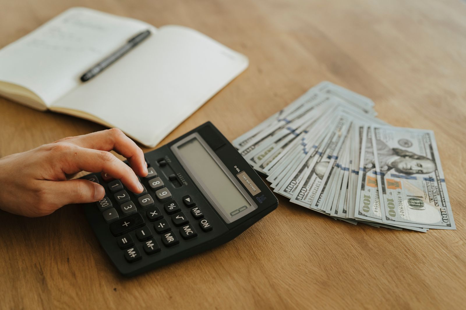 A hand using a calculator beside US dollar bills and an open planning notebook
